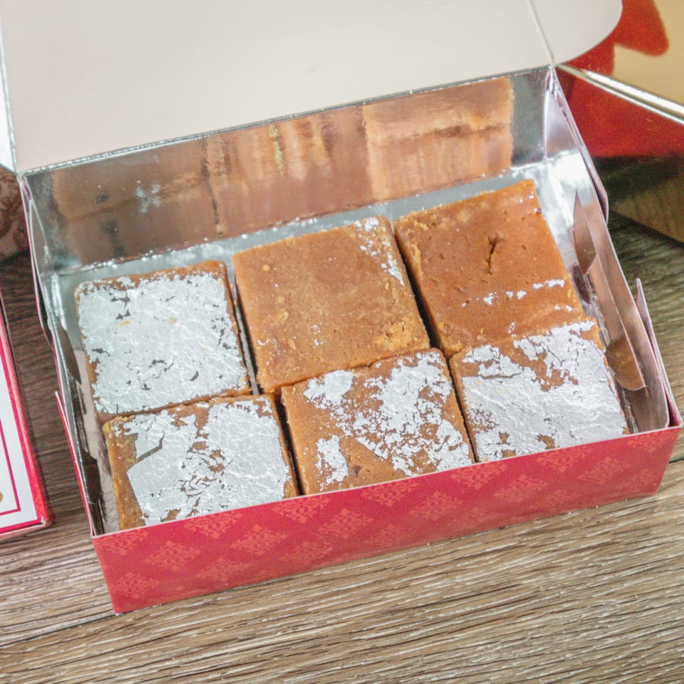 Assorted square Indian sweets, some dusted with edible silver leaf, arranged in an open red gift box.