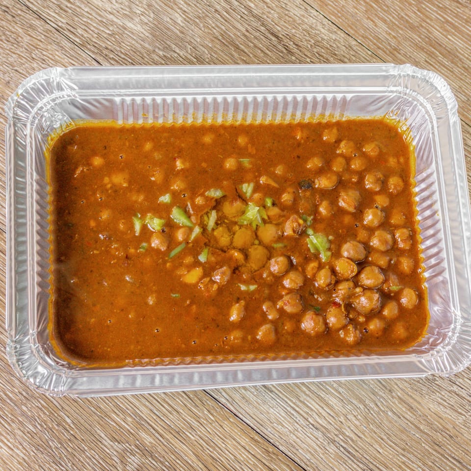 Aluminum tray filled with chickpeas in thick brown curry, garnished with chopped green peppers on a wooden table.