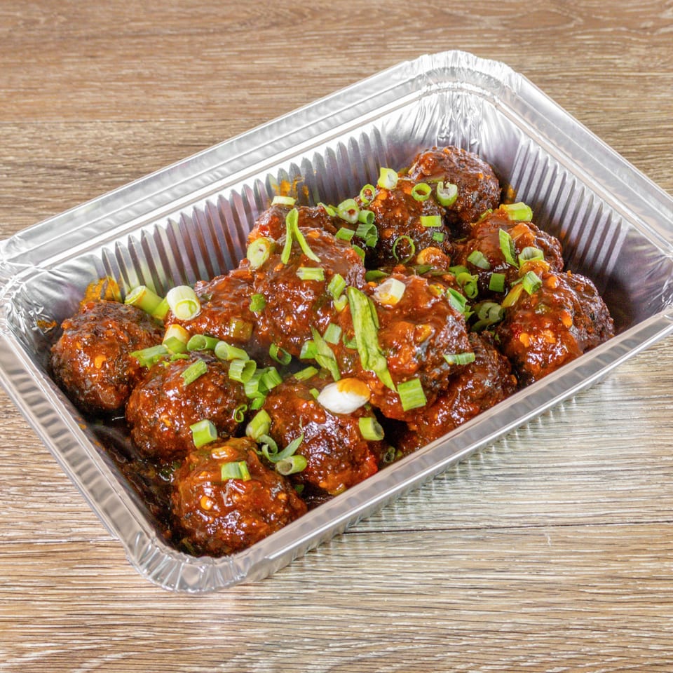 Glazed meatballs topped with sliced green onions fill a disposable aluminum tray on a wooden table.