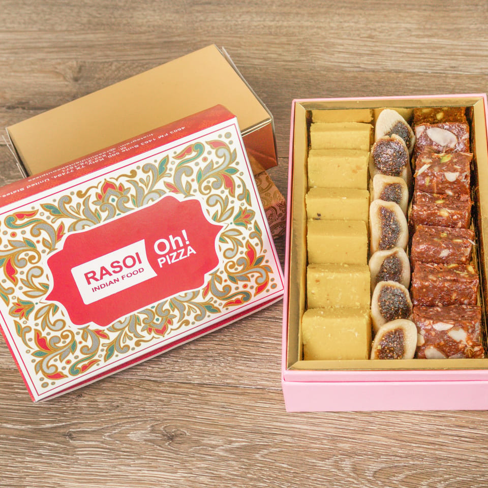 Rasoi Indian Food gift box displaying assorted colorful sweets beside its ornate branded packaging on a wooden table.