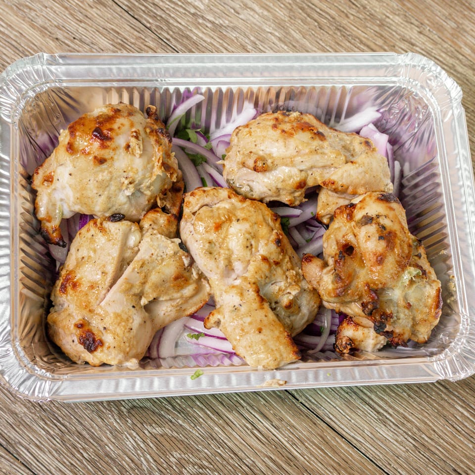 Grilled chicken pieces served in a foil tray over sliced red onions and fresh herbs.