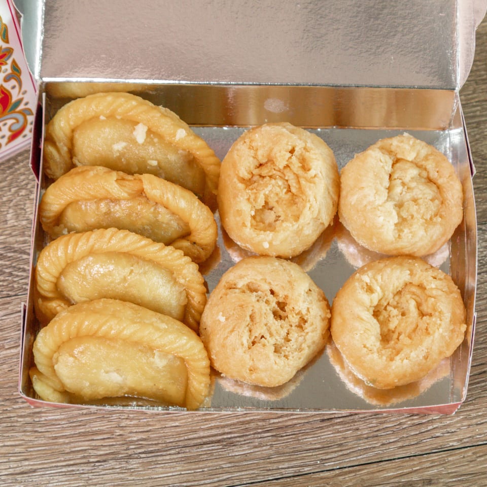 Open box of golden pastries, including crescent-shaped sweets and crumbly round cookies, on a wooden table.