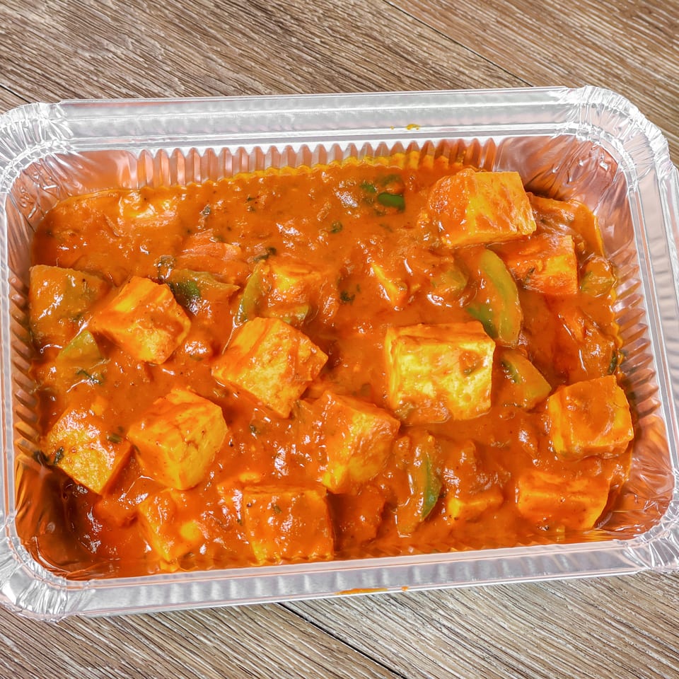 Paneer tikka with green peppers in a creamy orange sauce, served in a rectangular foil container on a wooden table.
