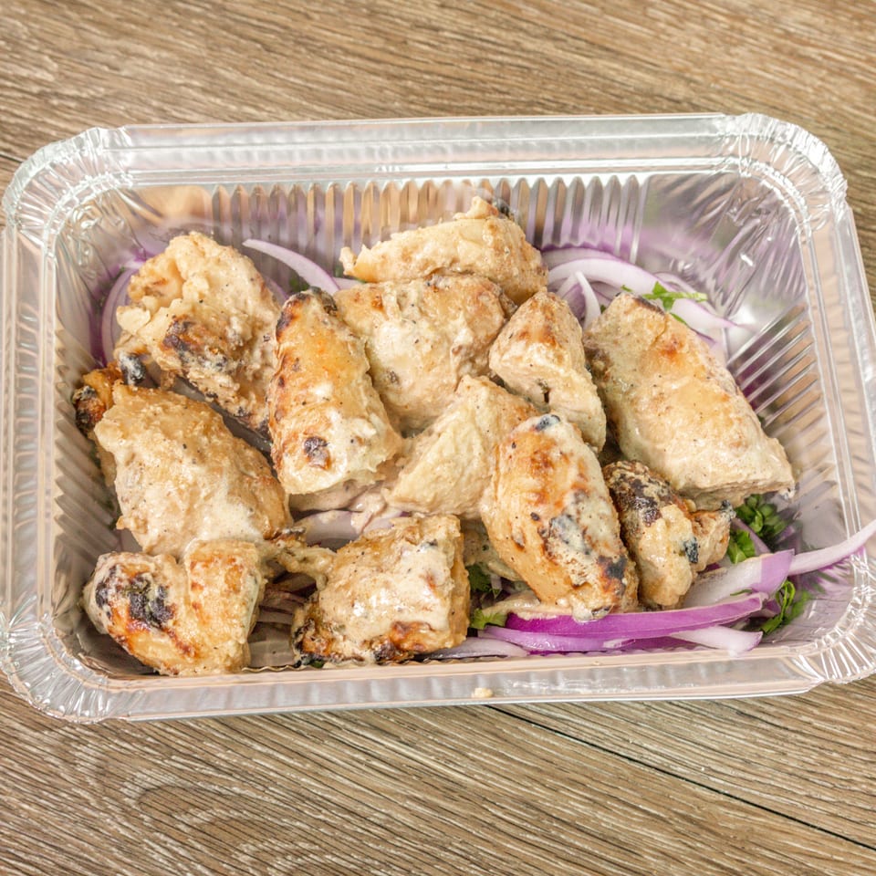 Grilled chicken pieces with charred edges, red onion slices, and herbs in a foil takeout tray.
