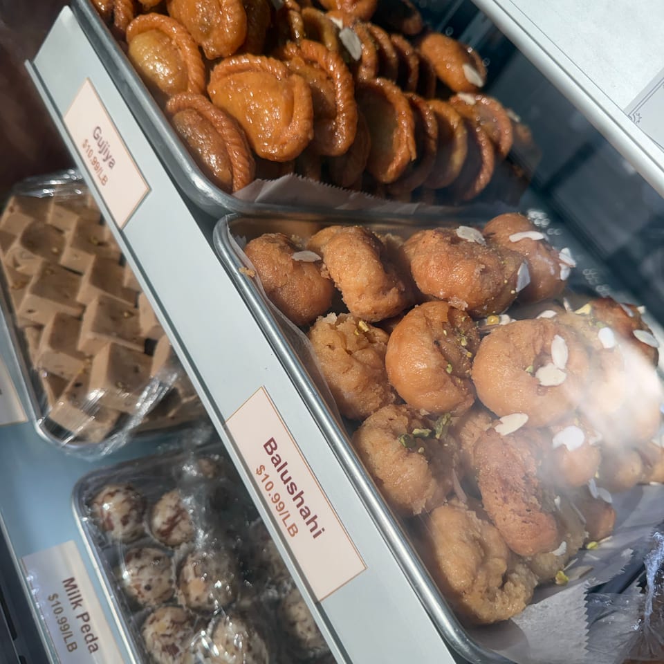 Indian sweets, including syrup-glazed gujiya and balushahi pastries, arranged in trays inside a bakery display case.