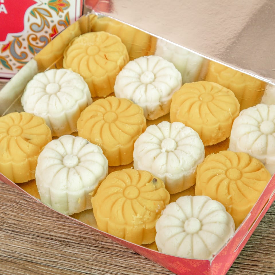 Assorted yellow and white flower-shaped mooncakes arranged in a decorative red gift box on a wooden table.
