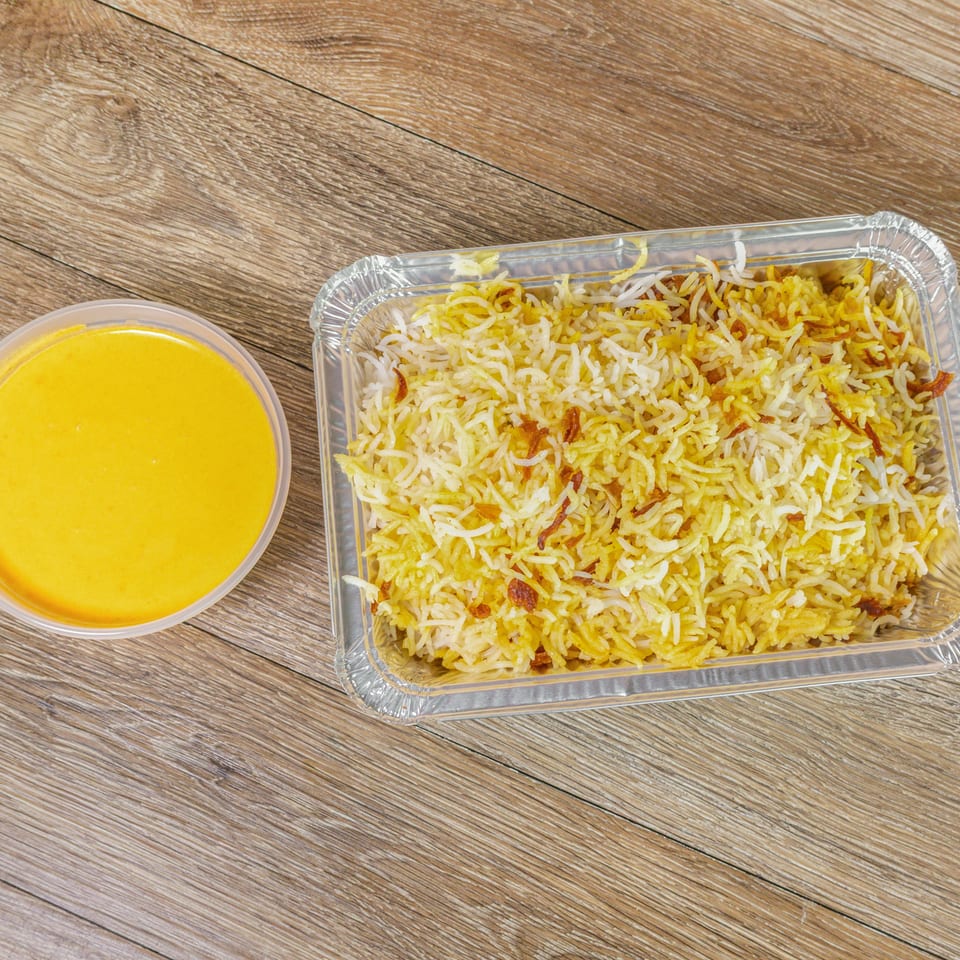 Aluminum tray of biryani rice with fried onions beside a bowl of yellow curry on a wooden table.