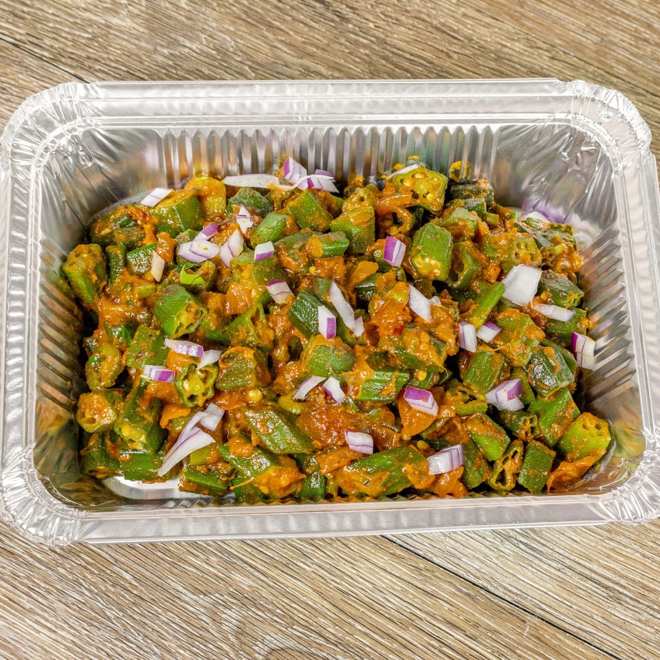 Spiced chopped okra garnished with red onion in a foil tray on a wooden table.