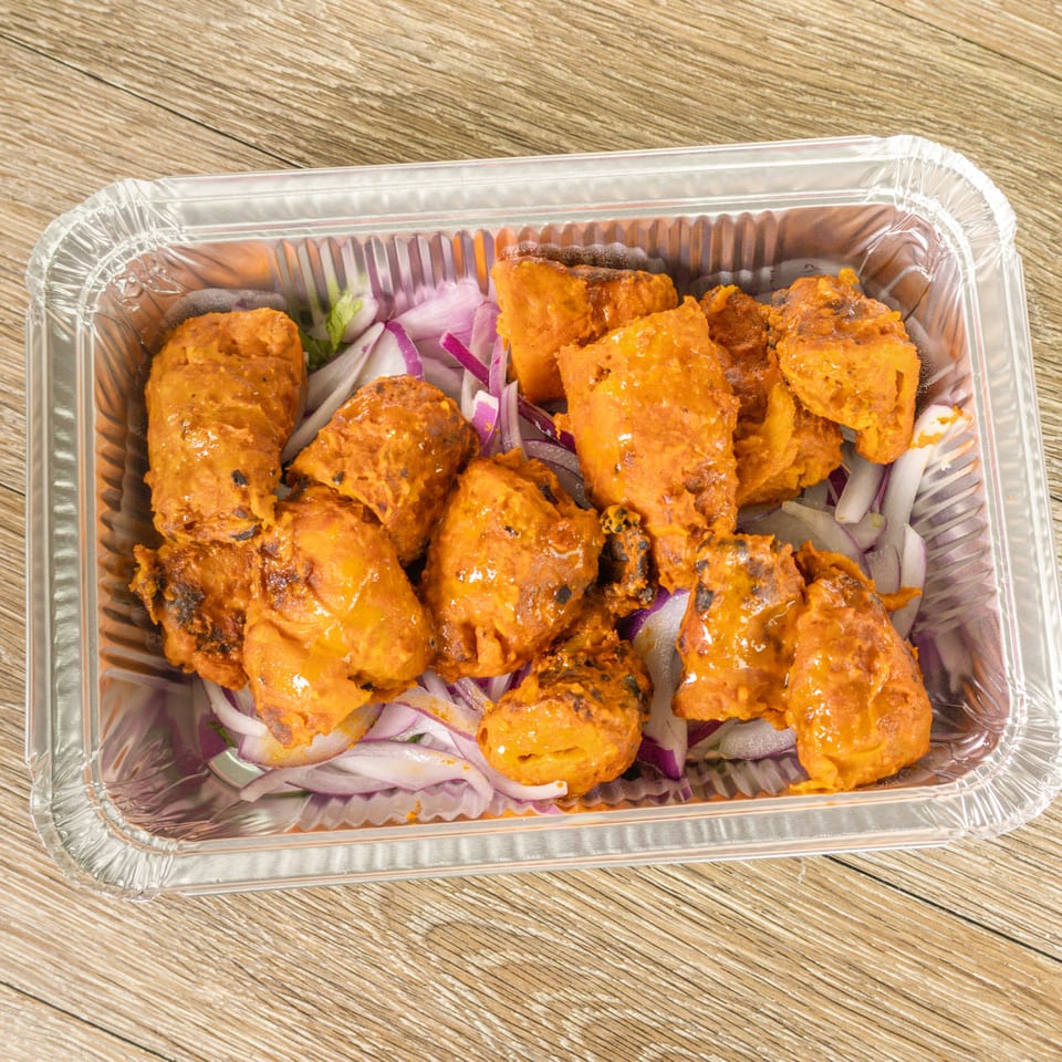 Golden-orange grilled chicken pieces served with sliced red onions in a disposable aluminum tray on a wooden table.